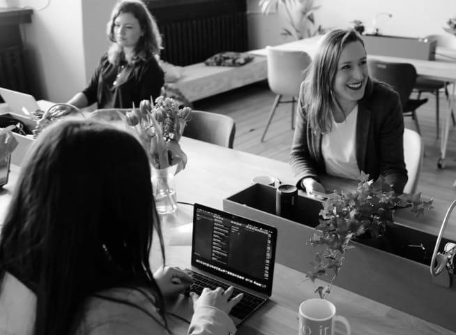 Three woman are sitting around a table. Two of them are working on computer while one of them is laughing.There are also coffee mug, flowers and other working material on the table.